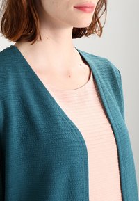 Tyrkysový texturovaný kardigan s otevřeným předním dílem, vrstvený přes světle růžový ribbovaný top. Zaměření na detaily tkaniny a barevný kontrast.