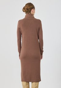 Rochie tricotată maro cu guler înalt, mâneci lungi și o siluetă ajustată. Prezintă o textură striată și slituri laterale la tiv.