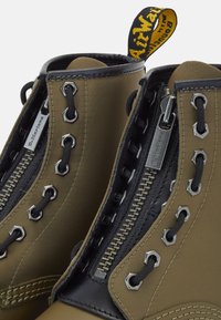 Gros plan sur des bottes Dr. Martens vert olive avec des lacets noirs, des œillets argentés et des fermetures éclair centrales avec des tirettes de fermeture éclair marquées.
