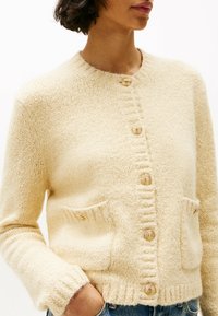 Crème-kleurige cardigan van textuur gebreid materiaal, met een ronde halslijn, gouden knopen en twee frontzakken.