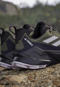 Zapatos de senderismo Terrex, con una parte superior de malla verde oscuro, acentos negros, suela de goma texturizada y tres franjas blancas distintivas a los lados.