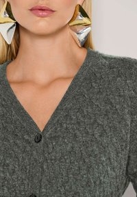 Frau trägt große geometrische Ohrringe in Gold- und Silbertönen, mit einer grauen, kabelgestrickten V-Ausschnitt-Charité, die mit schwarzen Knöpfen geschlossen ist.