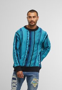 Uomo con capelli corti e barba, che indossa un maglione blu a fantasia e jeans strappati con toppe colorate, in piedi davanti a uno sfondo semplice.