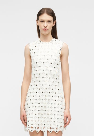 Γυναίκα που φοράει λευκό δαντελωτό φόρεμα χωρίς μανίκια με κυκλικές κοψίματα και αντανακλαστικές διακοσμήσεις, στέκεται μπροστά σε ένα απλό φωτεινό φόντο.