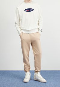 Ljusgräddfärgad sweatshirt med blå och röd "NIKE"-logotyp, kombinerad med beige joggers. Bärs med vita högklackade sneakers. Enfärgad bakgrund.