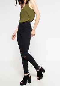 Oliivinvihreä v-kaula-aukollinen rento paita, yhdistettynä korkeavyötäröisiin mustiin kapealahkeisiin farkkuihin, joissa on polvireiät, ja mustiin korokepohjaisiin korkokenkiin.