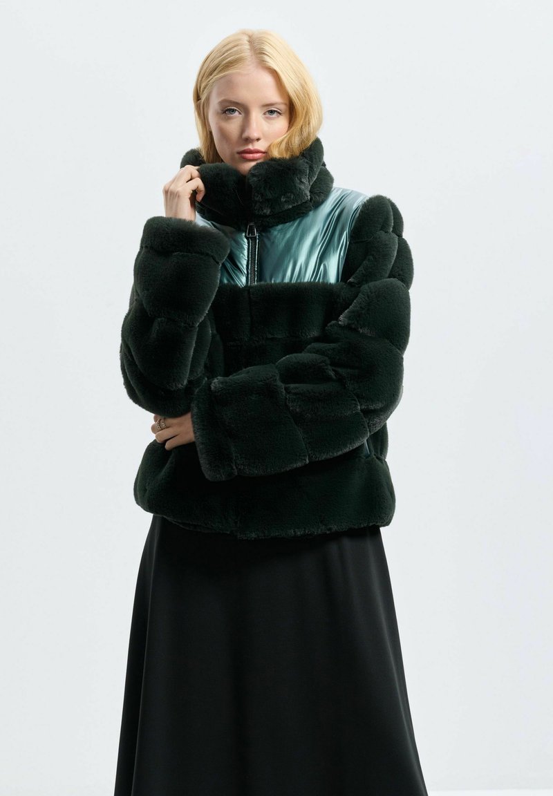 Giacca puffer in mimetica verde scuro in pelliccia sintetica con dettagli lucidi in teal; colletto alto, chiusura con zip e forma squadrata. Aspetto texturizzato e morbido.