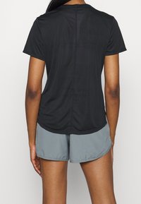 Persona vestida con una camiseta negra de manga corta y pantalones cortos deportivos grises, vista desde atrás contra un fondo blanco liso.