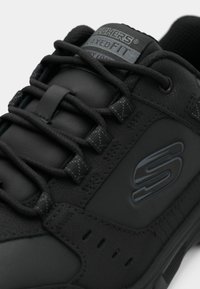 Črne atletske čevlje z gladkim sintetičnim zgornjim delom, teksturiranimi vezalkami, oblazinjenim ovratnikom in embosiranim logotipom na strani.