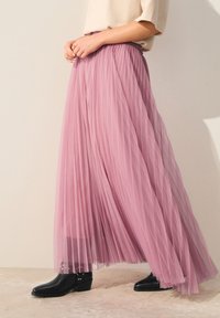 Jupe maxi plissée rose avec une texture transparente et une taille élastique, associée à des bottines noires avec des accents argentés.