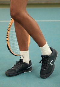 Svarta sportskor med texturerat material och vita accenter, parat med vita ankelsockor, placerade bredvid ett tennisracket på en blå bana.