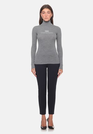 Donna in piedi che indossa un maglione grigio attillato con collo alto e pantaloni neri con tacchi alti neri su sfondo bianco.
