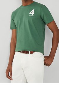 Camiseta de algodón verde con mangas cortas, que presenta un número "4" impreso en blanco y el logo "HACKETT" en el pecho. Combinada con pantalones blancos.