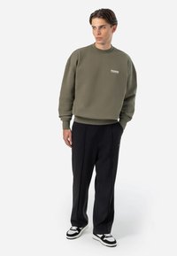 Olive green sweatshirt with ribbed cuffs and neckline, featuring white text logo. Black trousers with a straight fit and side seam detail.
