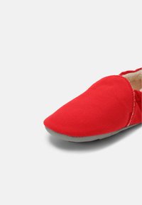 Czerwony canvasowy but wsuwany z okrągłym czubkiem, fakturowaną powierzchnią i szarym gumowym spodem; cechuje się minimalnym przeszyciem i brakiem widocznych elementów metalowych.