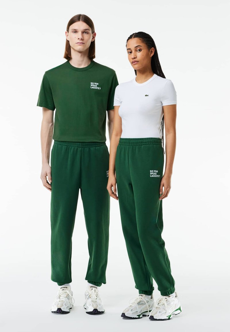 Zielone spodnie dresowe i dopasowana zielona koszulka z napisem; biała dopasowana koszulka z logo; buty sportowe z teksturowanym wzorem i akcentami.