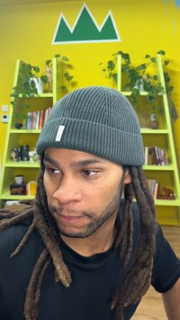 Dunkelgraue, gerippte Strickmütze mit einem kleinen weißen Etikett. Unter der Mütze sind Dreadlocks sichtbar. Im Hintergrund sind eine gelbe Wand und grüne Regale zu sehen.