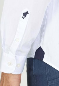 Chemise blanche à manches longues avec broderie de cheval marine sur le poignet, dotée d'une fermeture à boutons et d'un accent contrastant en marine à l'ourlet. Texture unie.