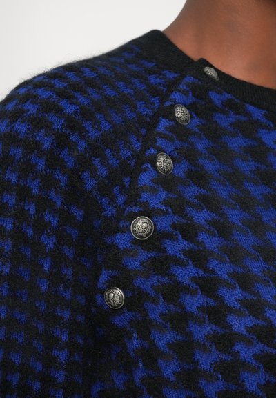Kootud sviiter sinise ja musta houndstooth mustriga, millel on ümmargune kaelus ja dekoratiivsed hõbedased nööbid õlal.