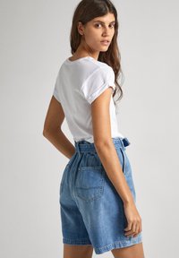 Sieviete ar gariem brūniem matiem, tērpusies baltā T-kreklā un augstās jostasvietas zilo džinsu šortos, stāv un skatās pār plecu.