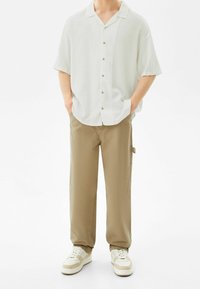 Camicia bianca a maniche corte con bottoni, pantaloni beige con tasca laterale e sneakers crema con dettagli beige, presentati su uno sfondo neutro.
