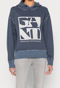 Mörkblå hoodie med en stor grafisk design i vit text. Mjuk tyg, luvtröja med dragsko, ribbade ärmslut och nederkant. Tillsammans med ljusgrå jeans.