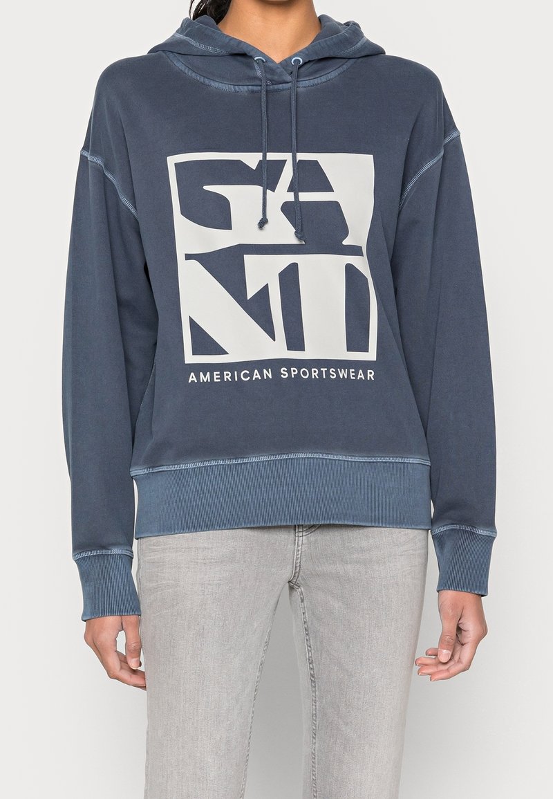 Mörkblå hoodie med en stor grafisk design i vit text. Mjuk tyg, luvtröja med dragsko, ribbade ärmslut och nederkant. Tillsammans med ljusgrå jeans.