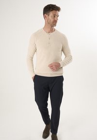 Beige gerippter Pullover mit langen Ärmeln und Drei-Knopf-Leiste, kombiniert mit dunkelblauen Hosen und braunen Wildleder-Stiefeletten.