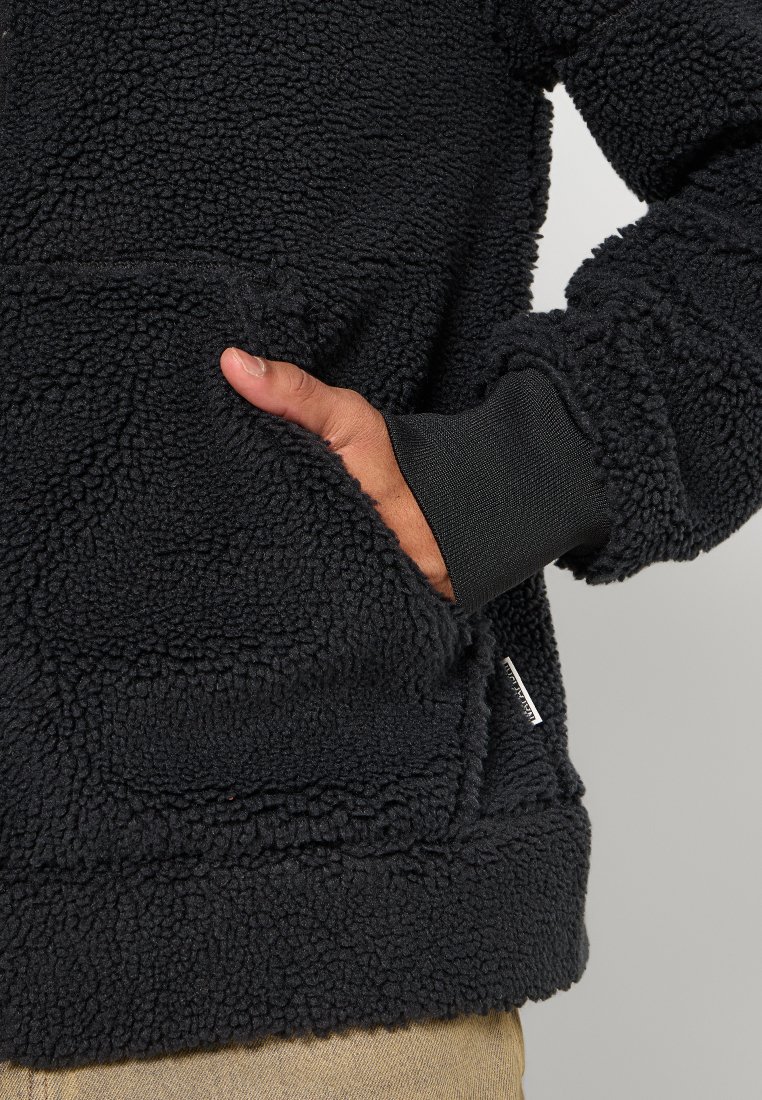 Schwarze Fleecejacke mit einer strukturierten, flauschigen Oberfläche, ausgestattet mit einer Fronttasche und einem gerippten Bündchen. Der Stoff ist weich und warm.