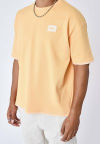 Camicia testurizzata gialla chiara a maniche corte con scollo rotondo e una piccola toppa con logo. Le cuciture bianche accentuano l'orlo e le maniche.