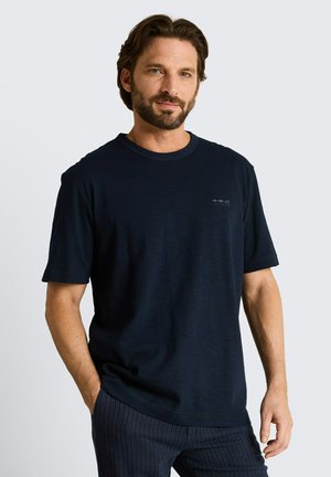 Mężczyzna z brodą ubrany w jednolity, ciemnogranatowy t-shirt i ciemne, w paski spodnie, stojący z jedną ręką w kieszeni.