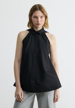 Žena v černém topu bez rukávů s límečkem a výřezy na ramenou a ve světlých šedých kalhotách stojící před jednobarevným bílým pozadím.