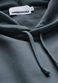 Donkergrijze hoodie met een buidelzak, trekkoorden en een zachte, gestructureerde binnenkant. Label met de tekst "ARMEDANGELS" in zwarte letters.