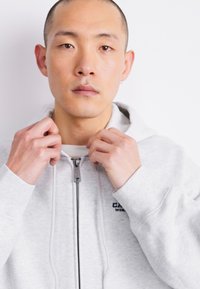 Light grey zip-up hoodie with a soft texture, featuring a hood and a small logo on the left chest. The model adjusts the zipper.