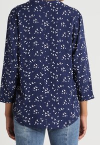 Navy blue blouse with three-quarter sleeves featuring a white bird pattern. Smooth fabric with a rounded hem and relaxed fit.