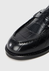 Zwarte leren loafer met een glanzende afwerking, voorzien van een krokodillenhuidpatroon, gestikte randen en een ronde neus.