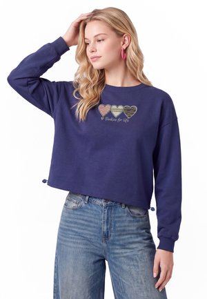 Ung kvinde med langt blondt hår iført en mørkeblå sweatshirt med tre mønstrede hjerter og tekst "Perfect for life", kombineret med blå jeans.