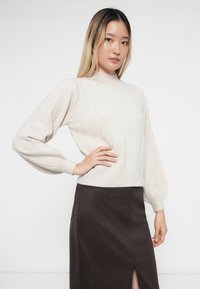Vaalean beige ribboitettu neule, jossa on korkea kaulus ja puffihihat, yhdistettynä tummanruskeaan hameeseen, jossa on sivureikä. Pehmeä rakenne ja istuva muotoilu.