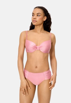 Mujer joven con cabello largo y rizado, vistiendo un top de bikini rosa brillante con doble tirante y pantalones de bikini de cintura alta, mirando hacia la izquierda.