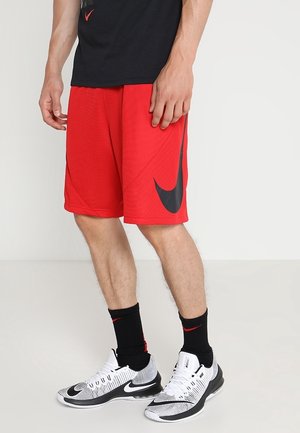Mand iført røde Nike basketballshorts, sorte Nike-strømper og hvide og sorte Nike-sneakers, stående på en ensfarvet lys baggrund.