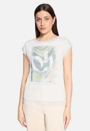 Vrouw met steil donker haar, gekleed in een wit topje met korte mouwen met een abstract blauw, groen en wit ontwerp en beige broek, staand tegen een effen achtergrond.