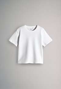 Lihtne valge lühikese varrukaga ümara kaelusega särk ripub helehalli seina taustal.