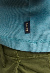 Una tela de ribete azul claro cerca del dobladillo presenta una etiqueta azul oscura con "Superdry" en letras doradas; combinada con pantalones verde oliva.