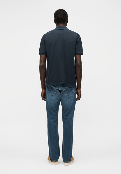 Άνδρας που στέκεται με την πλάτη, φοράει σκούρο κοντομάνικο πόλο μπλουζάκι, μπλε τζιν και καφέ παπούτσια σε απλό φωτεινό φόντο.