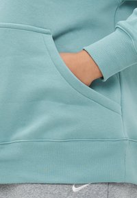 Mão repousada dentro do bolso da frente de um sweatshirt azul-água com punho canelado, acima da cintura de calças de treino cinzentas da Nike.