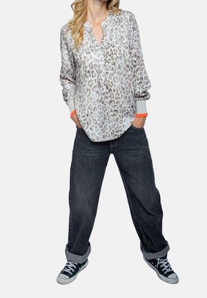 Person trägt eine langärmelige Bluse mit Leopardenmuster, lockere dunkle Jeans mit Umschlägen und schwarze flache Sneakers, steht mit den Händen in den Taschen.