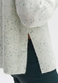 Weißer strukturierter Pullover mit feinen blauen und schwarzen Sprenkeln, ausgestattet mit einem gerippten Design und seitlichen Schlitzen. Getragen über dunkelgrünen Hosen.
