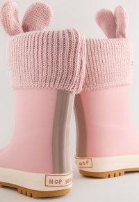 Rosa Regenstiefel aus Gummi mit einem gestrickten Bündchen und Hasenohren, strukturierter Oberfläche und einer beigen Sohle, auf der hinten "HOP HOP" aufgedruckt ist.