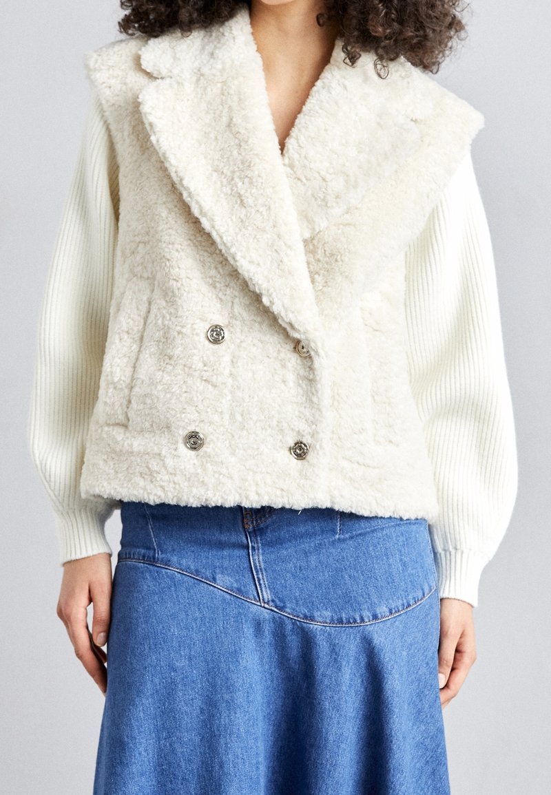 Gilet in pelliccia sintetica bianca con design doppiopetto, bottoni argentati e tasche laterali. Indossato sopra un top bianco a maniche lunghe a coste.