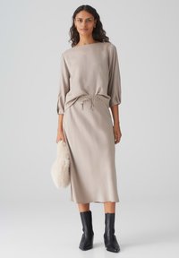 Beige, satinähnliches Kleid mit Boot-Ausschnitt, elastischer Taille und dreiviertellangen Ärmeln. Kombiniert mit schwarzen Stiefeletten und einer flauschigen weißen Clutch.